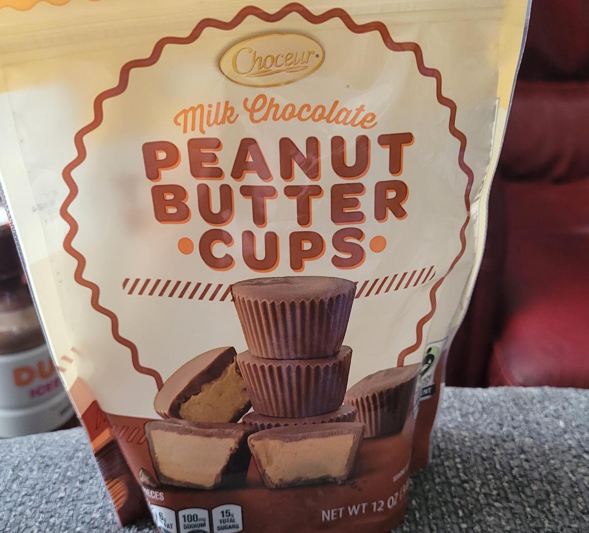 A bag of Choceur Milk Chocolate Peanut Butter Cups is shown, featuring an image of stacked chocolate cups on the front. The bag is 12 ounces and is placed on a gray surface.