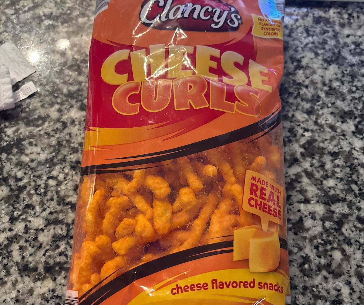 A bag of Clancy's Cheese Curls sits on a granite countertop. The packaging is orange and yellow, featuring images of cheese and highlighting that the snacks are "made with real cheese.