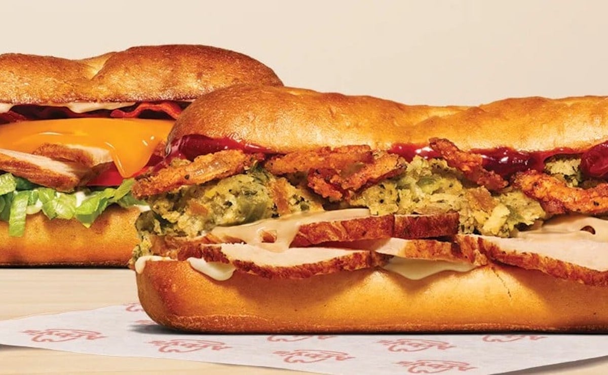 Two toasted sub sandwiches, one filled with lettuce, tomato, cheddar cheese, and deli meat, and the other with turkey, fried stuffing, and cranberry sauce, displayed on sandwich paper.