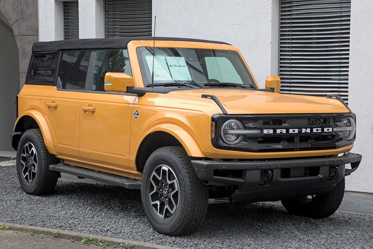 A yellow Ford Bronco SUV with black trim and a soft top is parked on a gravel surface in front of a white building with gray window blinds.