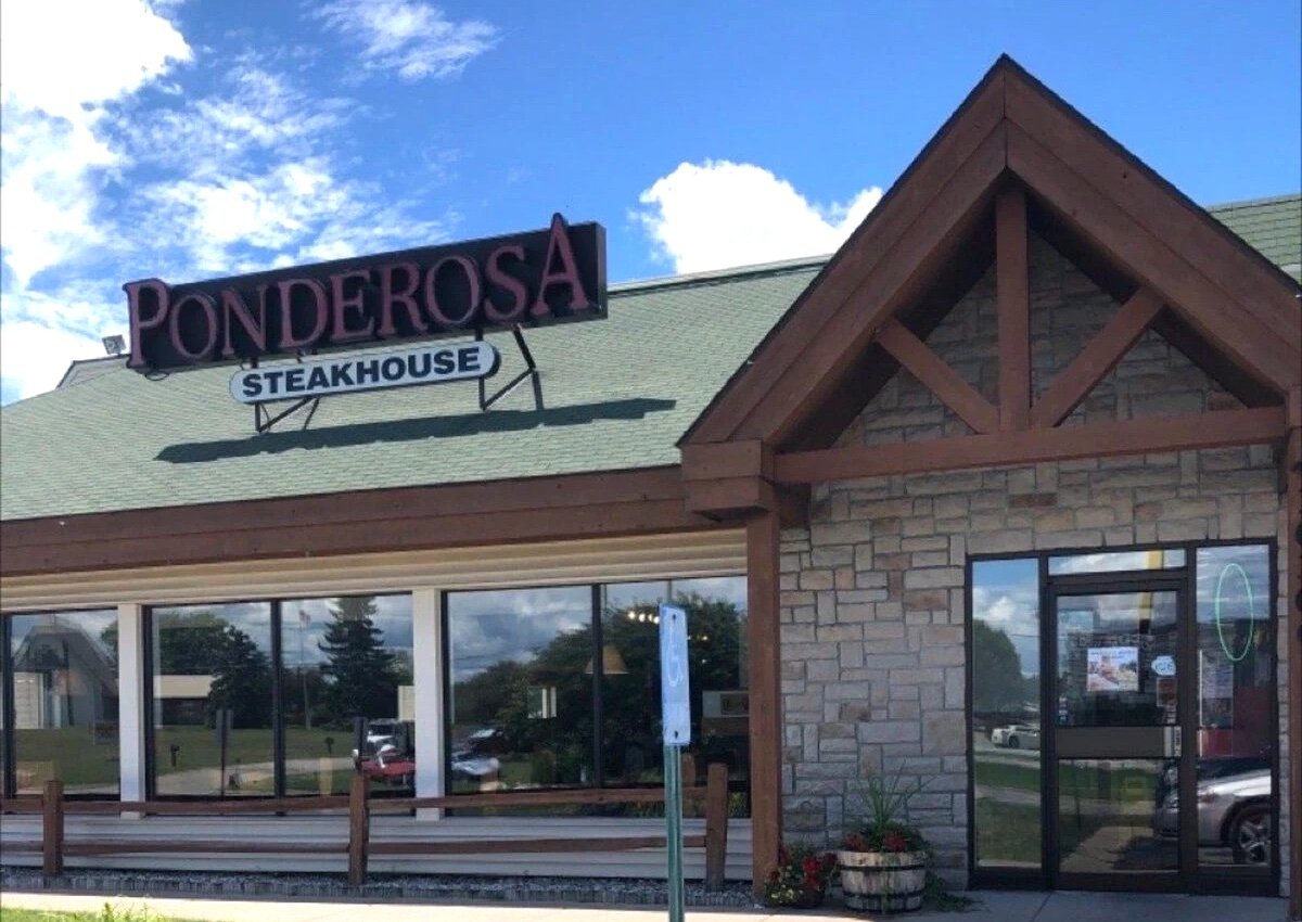 A Ponderosa Steakhouse restaurant with a stone and wood exterior, large windows, and a sign on the roof under a blue sky with scattered clouds.