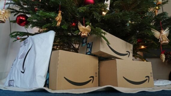 Several Amazon delivery boxes and a package are placed under a decorated Christmas tree with red baubles and straw ornaments.