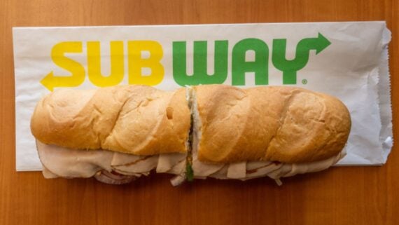 A Subway sandwich filled with deli meat sits on a white Subway-branded paper wrapper atop a wooden surface.