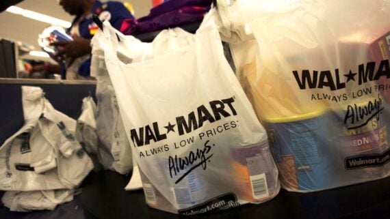Close-up of Walmart plastic shopping bags filled with groceries on a checkout counter, with a person in the background handling more bags. The bags are labeled “WAL-MART Always Low Prices. Always.”