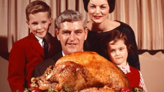 A family of four, dressed in formal clothing, smiles behind a large roasted turkey on a decorated platter, reflecting the joy and tradition of a Thanksgiving dinner as well as the evolving Thanksgiving dinner cost.