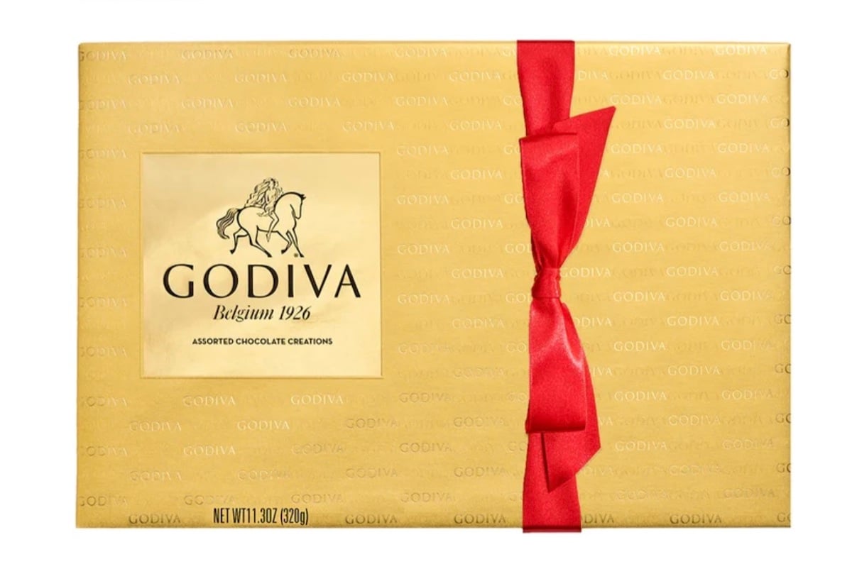 A gold Godiva chocolate box with a red ribbon tied on the right side. The box features the Godiva logo with a horse and rider, and text reading "Godiva Belgium 1926 Assorted Chocolate Creations.