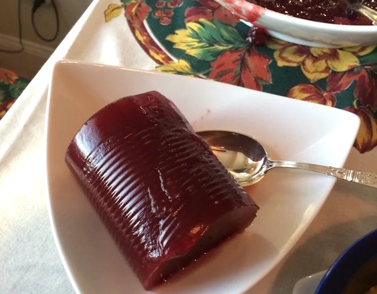 A cylindrical portion of canned cranberry sauce with visible can lines sits on a white triangular plate next to a silver spoon; a colorful autumn-themed tablecloth is in the background.