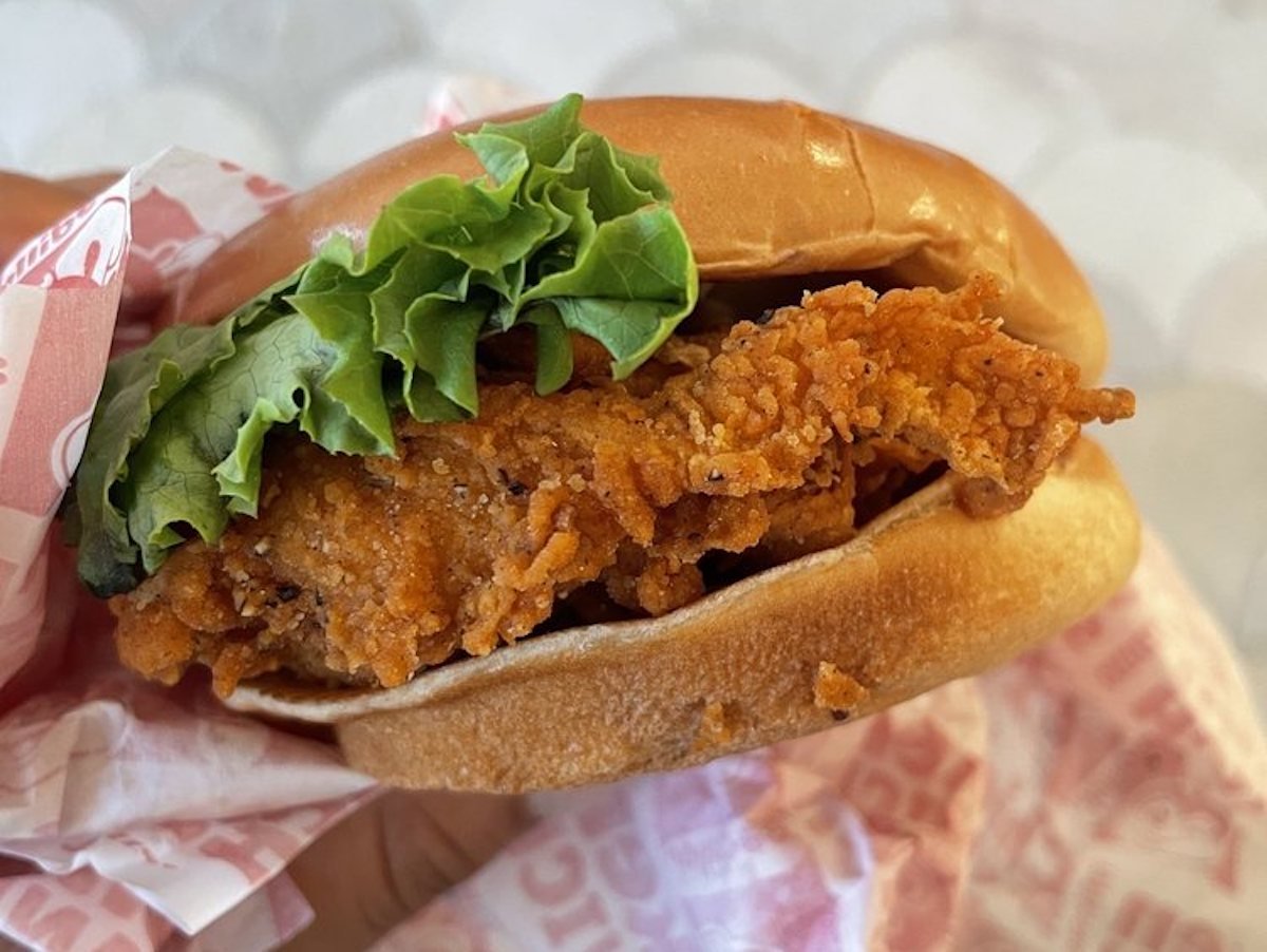 Close-up of a crispy fried chicken sandwich with leaf lettuce on a soft bun, partially wrapped in branded paper.