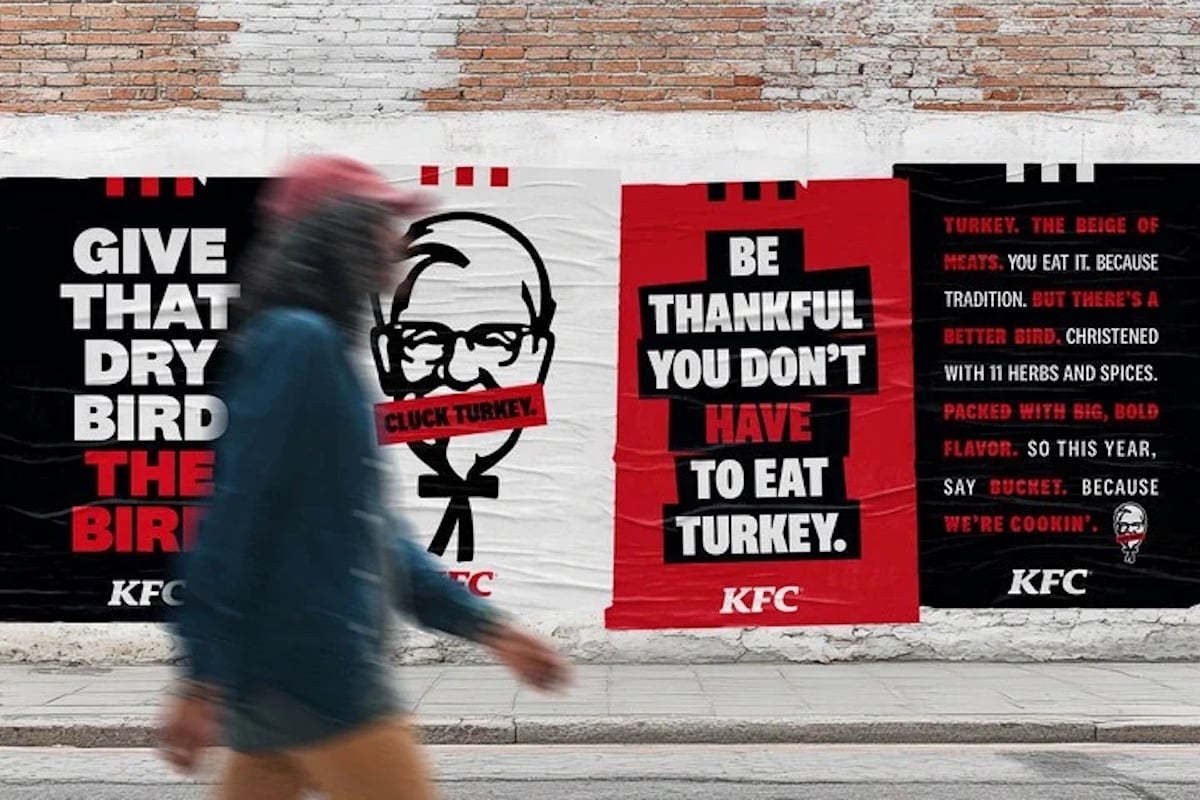 A person walks past posters on a brick wall advertising KFC with slogans like "Give that bird the bird" and "Be thankful you don’t have to eat turkey," featuring the KFC logo and red, black, and white colors.