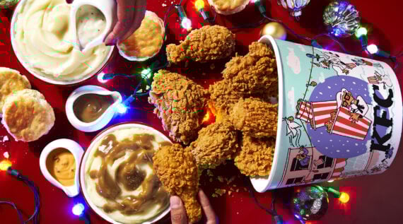 A festive table with a bucket of KFC fried chicken, mashed potatoes with gravy, biscuits, dipping sauces, and holiday string lights, all on a red background.