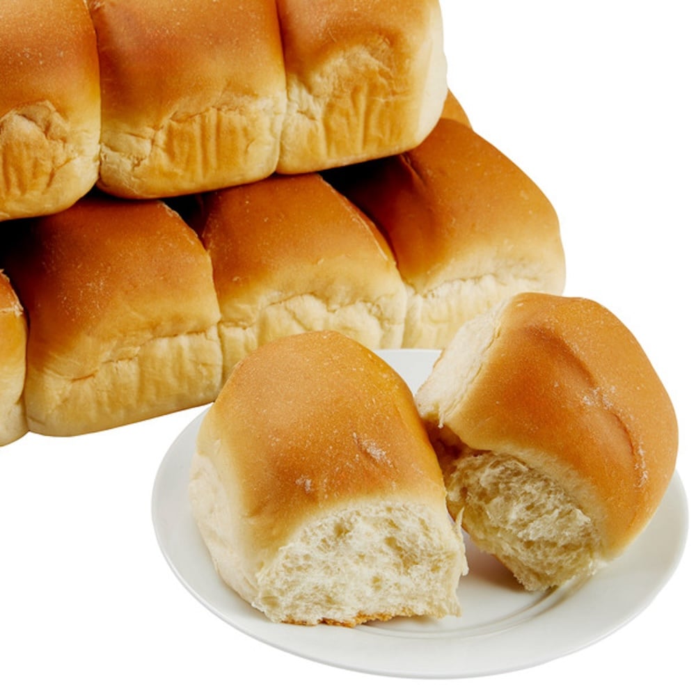A plate with two soft bread rolls, one partially torn open, sits in front of a stacked pile of similar golden brown rolls—just like the ones from Costco Thanksgiving spreads—on a white background.