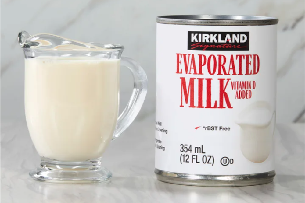 A glass cup filled with evaporated milk sits next to a can of Kirkland Signature evaporated milk, perfect for your Costco Thanksgiving recipes, labeled "Vitamin D Added" and "354 mL (12 FL OZ)," against a light background.