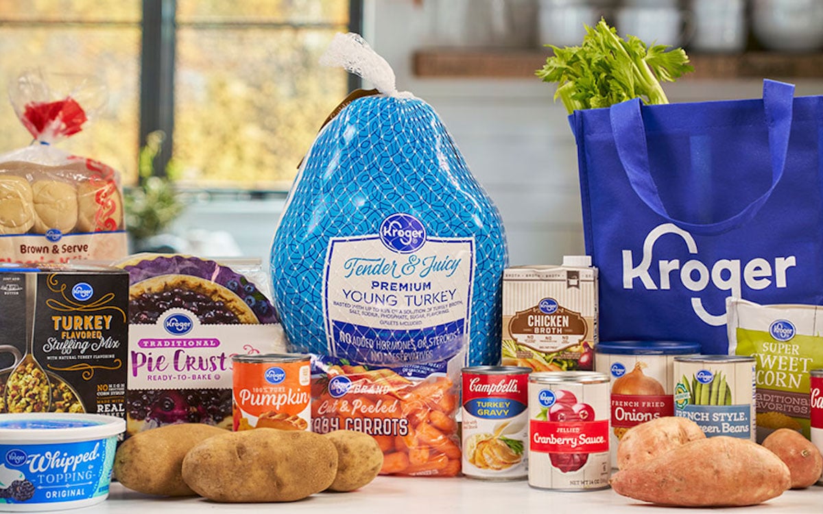 A variety of Kroger-branded Thanksgiving food items, including a frozen turkey, pie crust, canned vegetables, gravy, whipped topping, potatoes, celery, and a blue Kroger tote bag, displayed on a kitchen counter.
