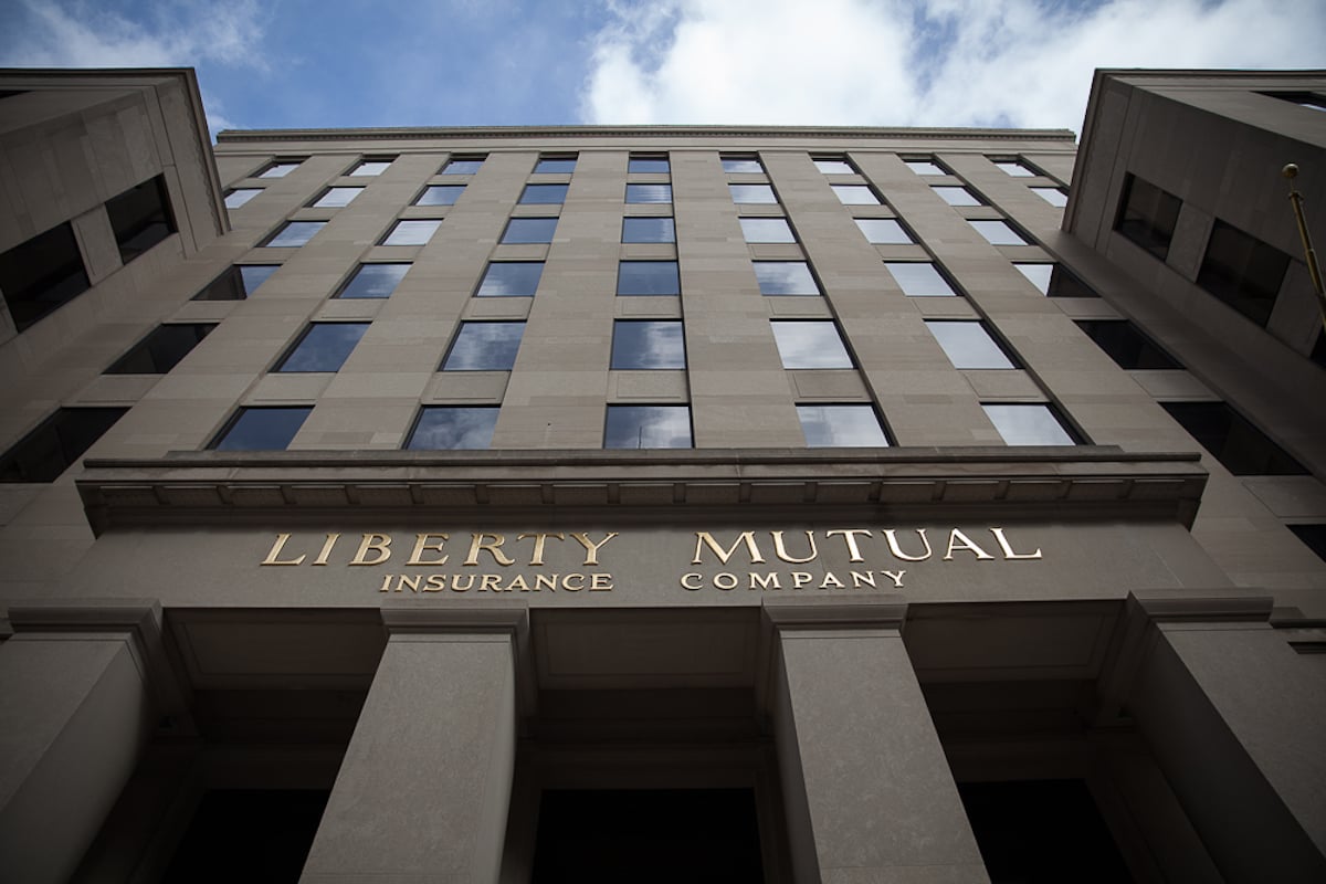 Looking up at the exterior of a tall office building with columns, featuring the gold-lettered sign “Liberty Mutual Insurance Company” above the entrance, set against a partly cloudy sky.