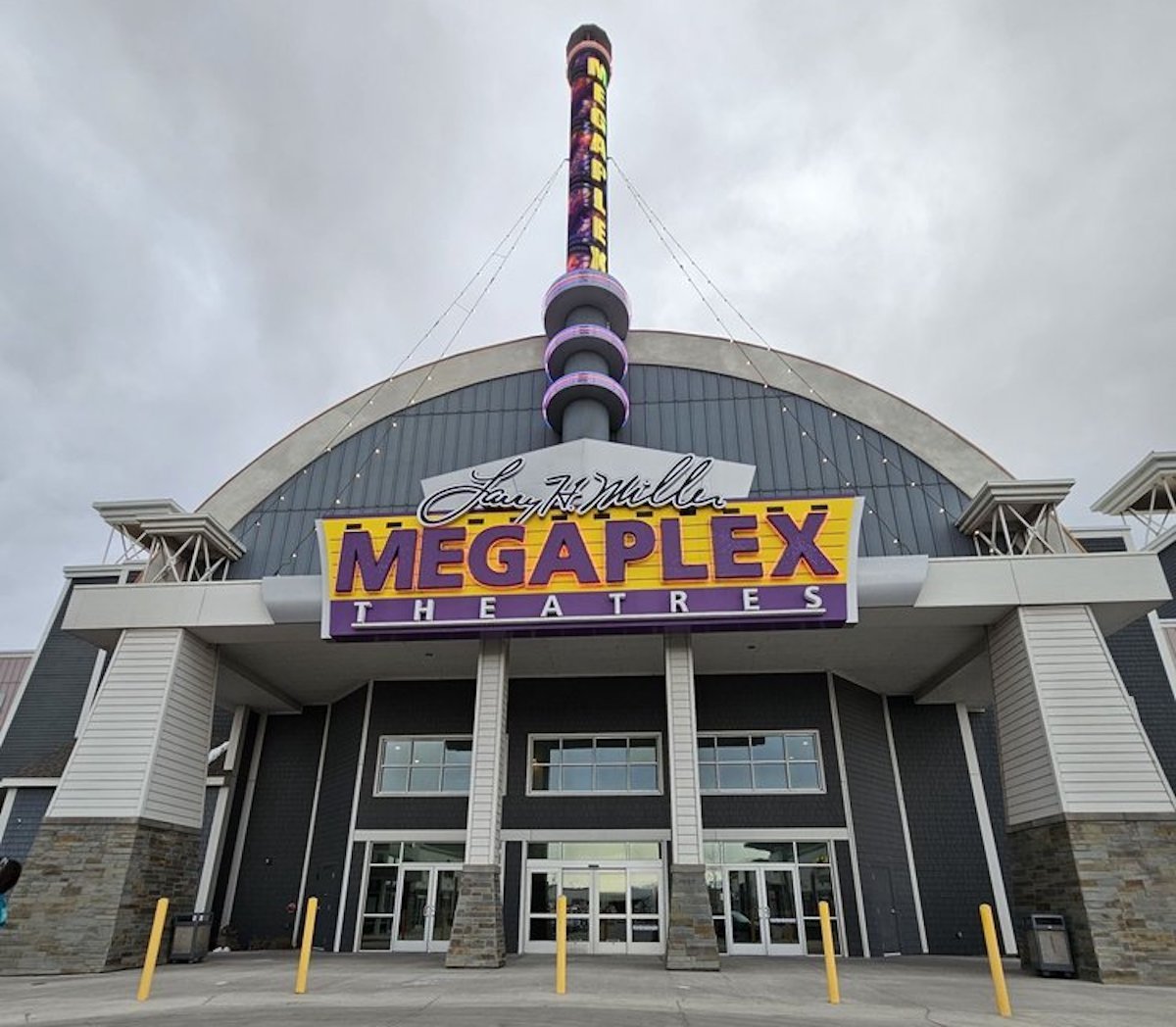 Exterior view of a Megaplex Theatres building with a large purple and yellow sign above the entrance and a tall illuminated marquee against a cloudy sky.