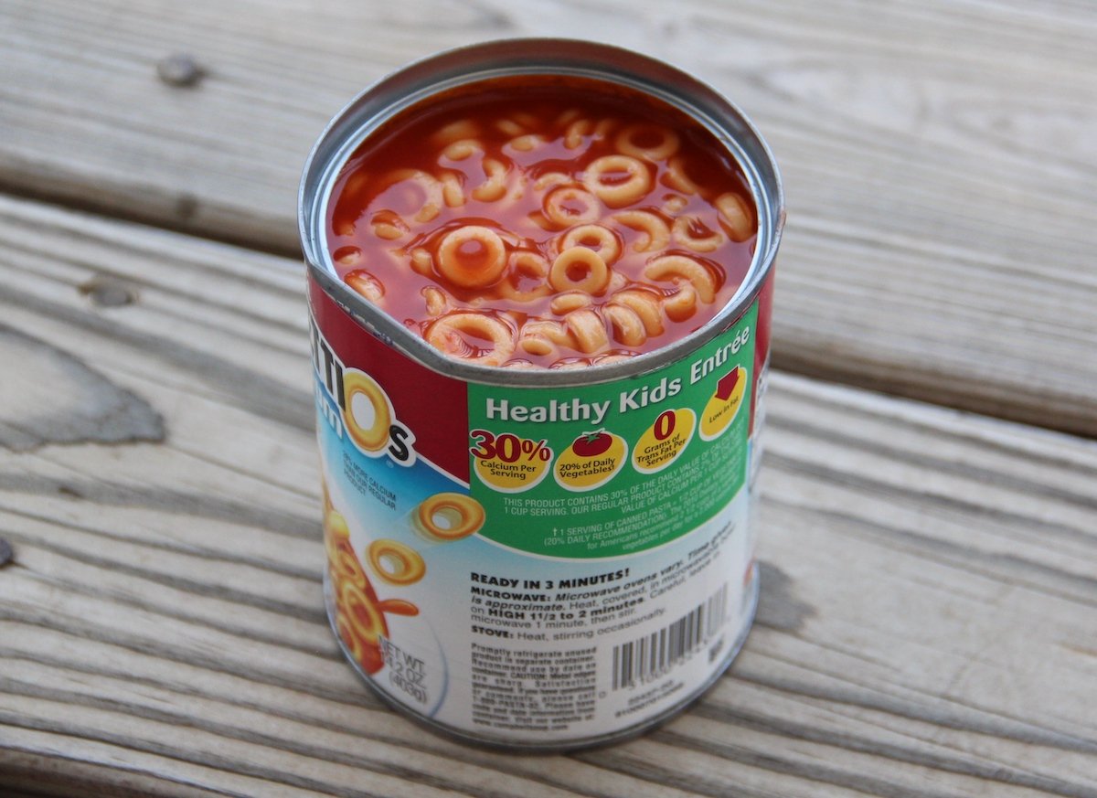 An open can of pasta rings in tomato sauce sits on a wooden surface. The label reads "Healthy Kids Entrée" and displays nutritional information. The pasta is visible at the top of the can.
