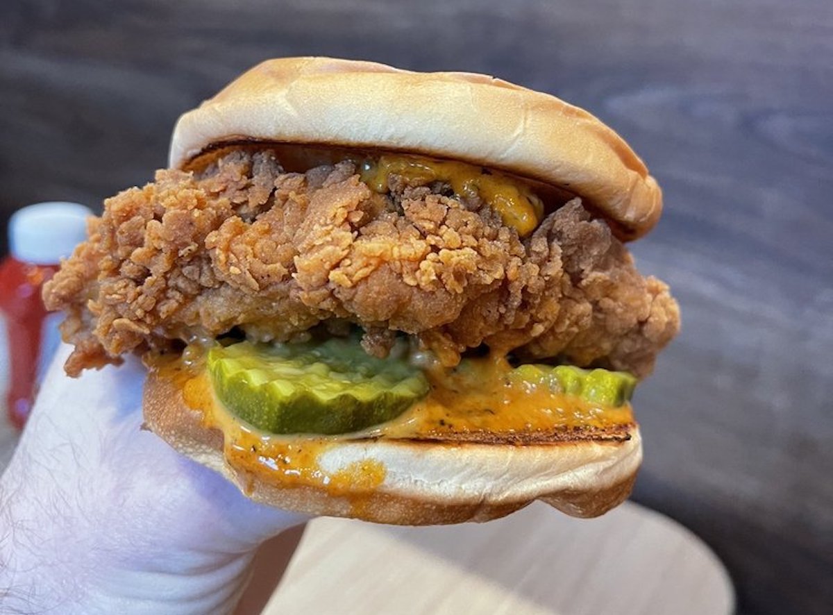 A hand holding a fried chicken sandwich with pickles and spicy orange sauce in a soft bun, against a blurred background.