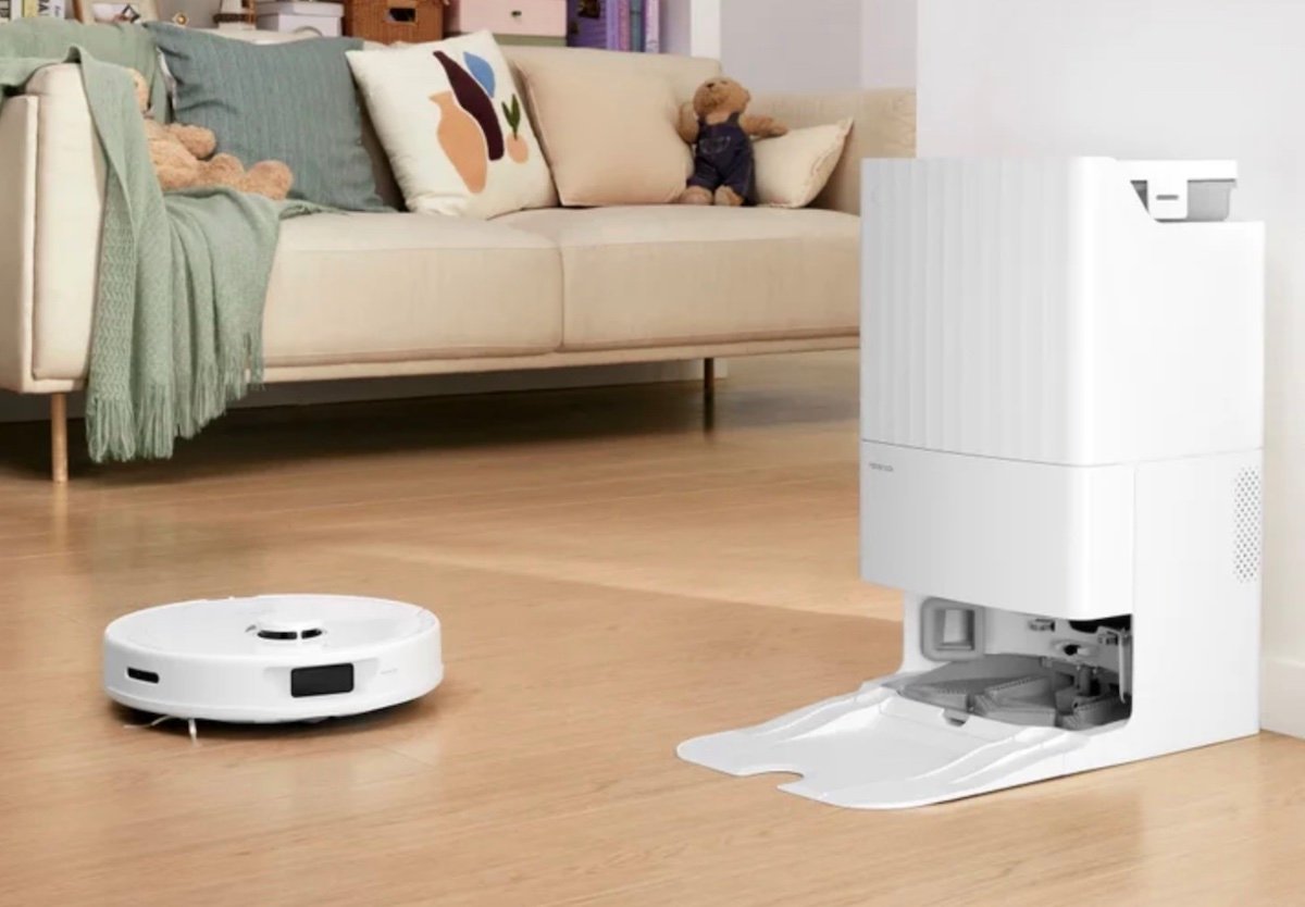 A white robot vacuum cleaner approaches its docking station on a wooden floor in a living room, with a beige sofa, green blanket, and teddy bear in the background.
