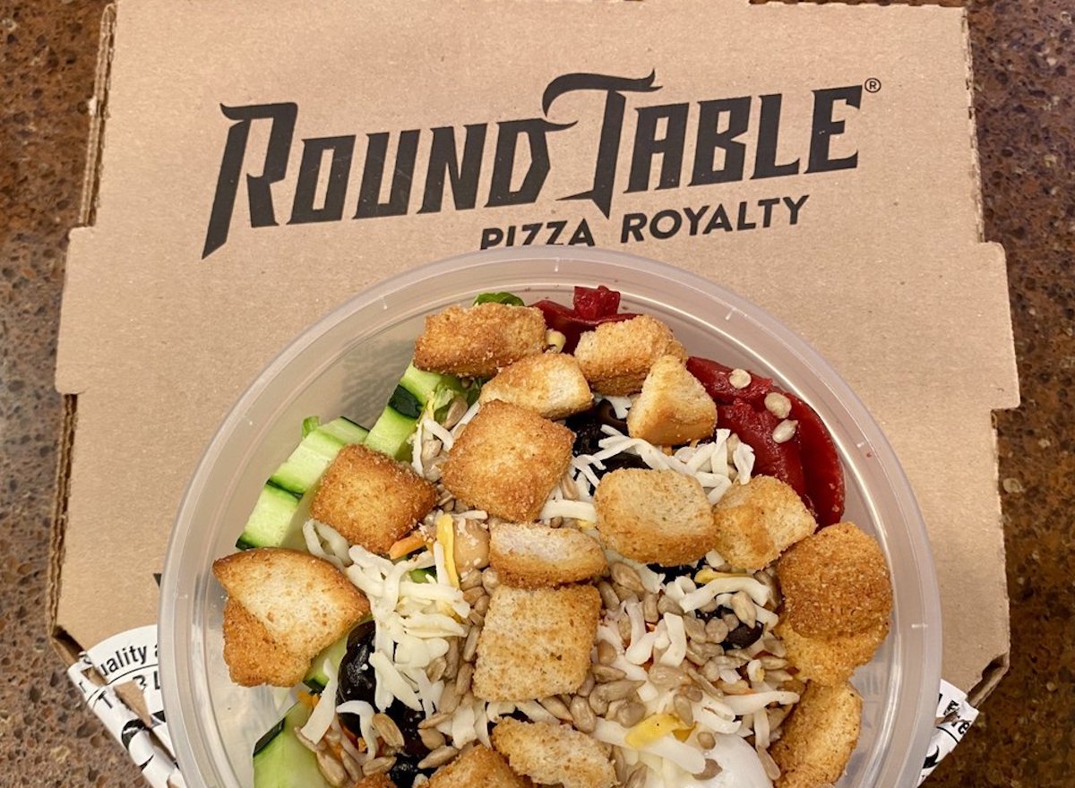 A plastic bowl of salad with croutons, shredded cheese, cucumber, and other toppings sits on top of a closed Round Table Pizza box.
