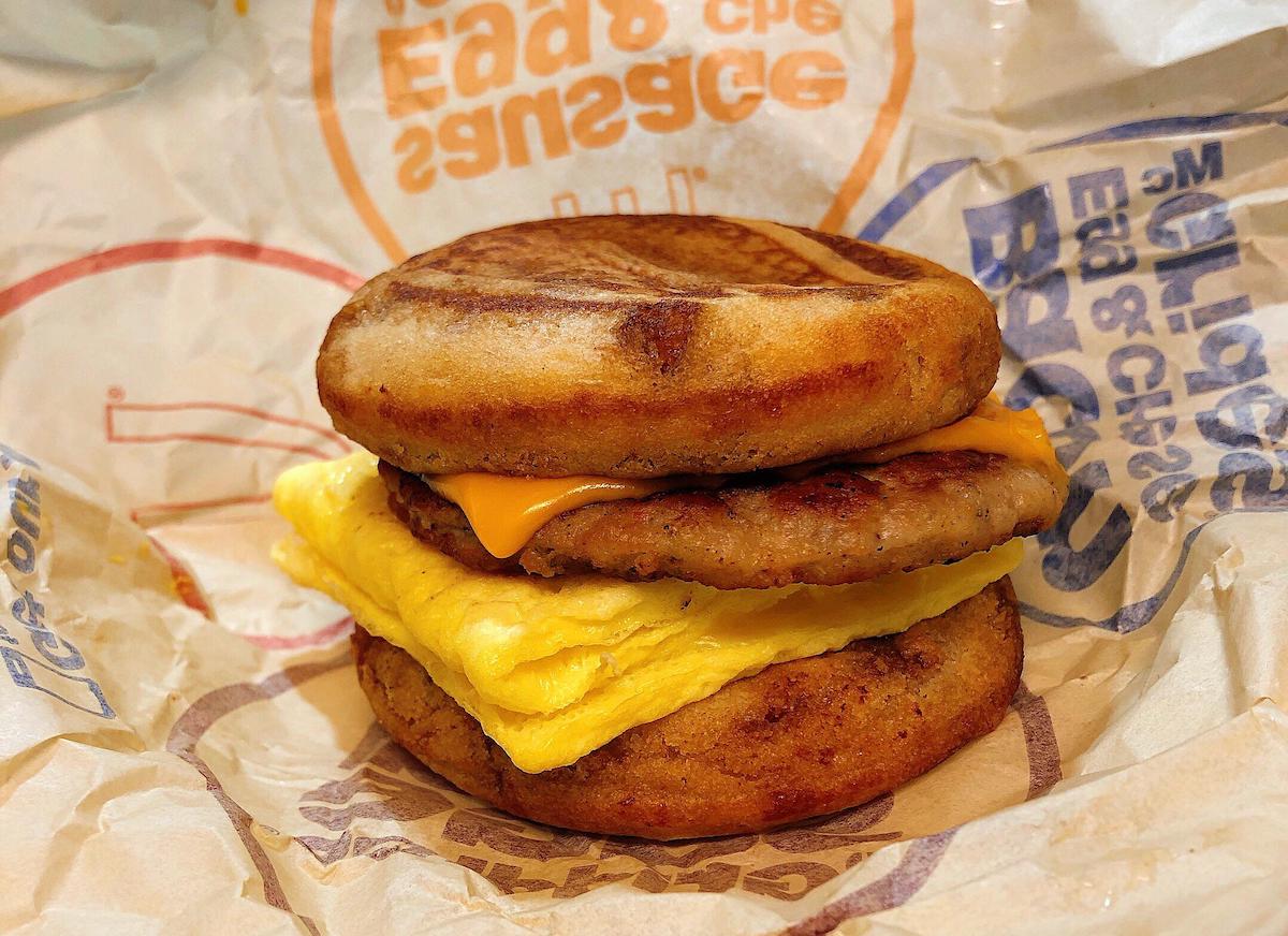 A breakfast sandwich with a folded egg, sausage patty, and melted cheese between two griddled pancake buns, wrapped in branded fast-food paper.