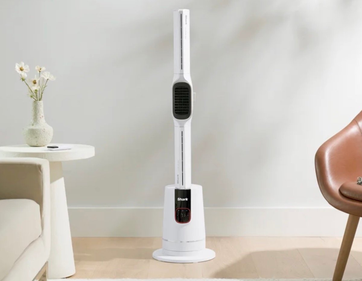 A tall, white bladeless tower fan stands on a light wooden floor between a beige sofa and a brown chair, with a small side table and vase of flowers nearby.