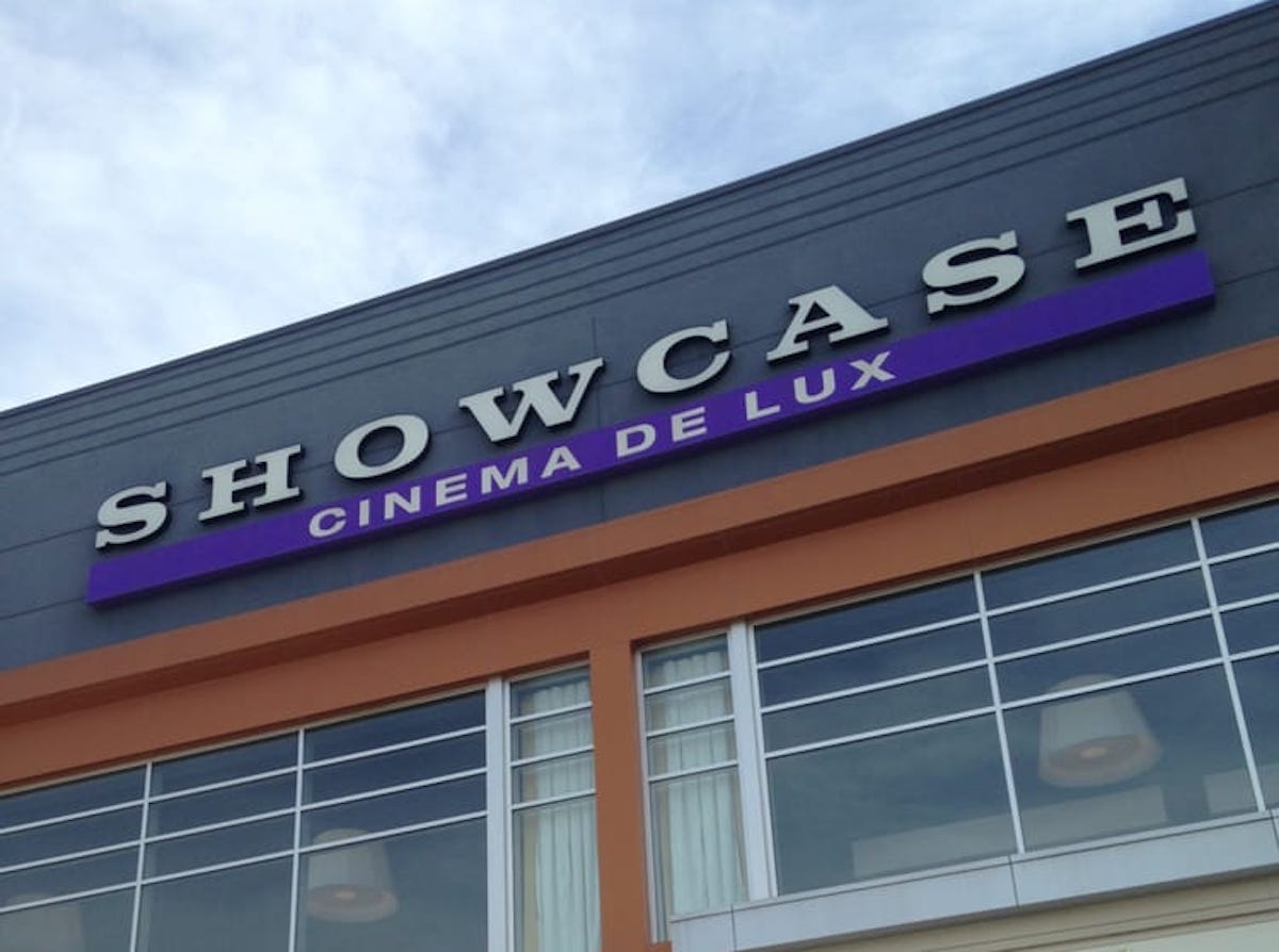 The exterior of a building with a sign that reads "SHOWCASE CINEMA DE LUX" in large letters against a gray wall above large windows.