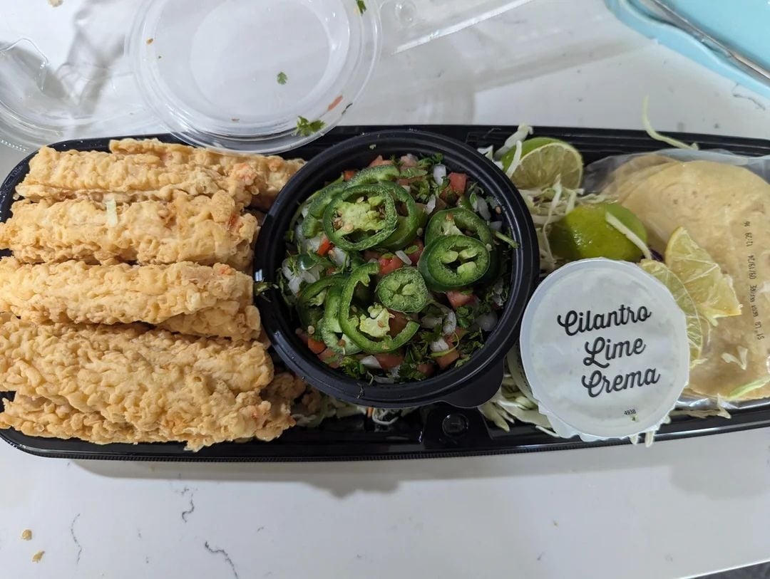 A takeout container with fried fish strips, a cup of pico de gallo topped with sliced jalapeños, lime wedges, mini tortillas, shredded cabbage, and a container labeled "Cilantro Lime Crema"—perfect for easy Costco dinners.