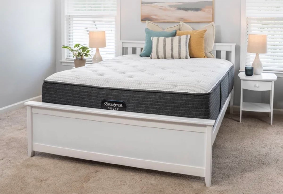 A neatly made bed with a thick mattress, several decorative pillows, white bed frame, and two nightstands with lamps, set in a bright, carpeted bedroom with light-colored walls and windows.