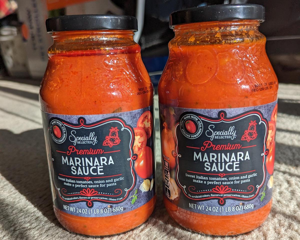 Two jars of "Specially Selected Premium Marinara Sauce" sit side by side on a beige surface, with sunlight highlighting their red tomato contents and black, red, and white labels.