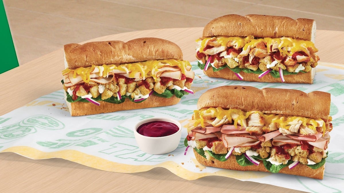 Three sub sandwiches filled with sliced turkey, ham, fried chicken, cheese, lettuce, onions, and sauce are placed on sandwich paper. A small cup of red dipping sauce sits in front of the subs on a wooden table.
