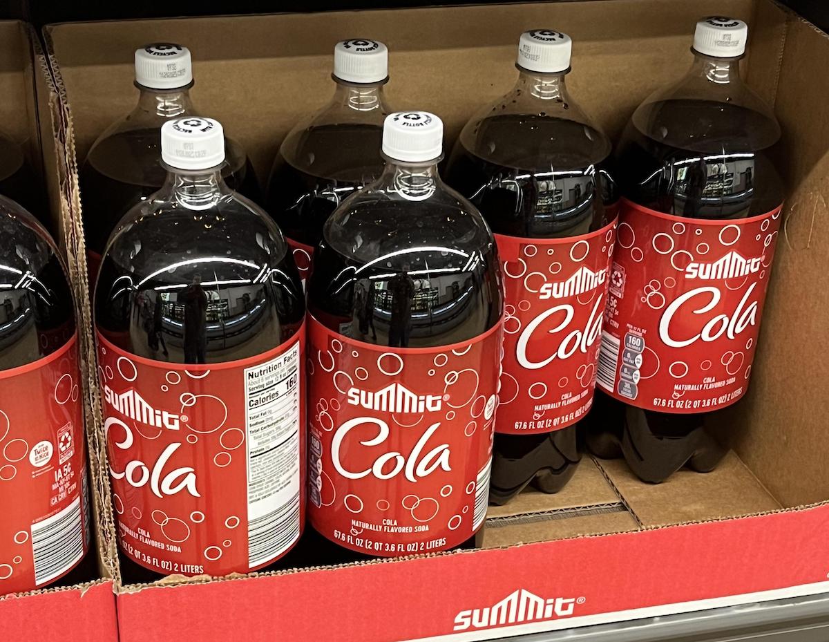 Several large bottles of Summit Cola with red labels and white text are displayed upright in a cardboard box on a store shelf.