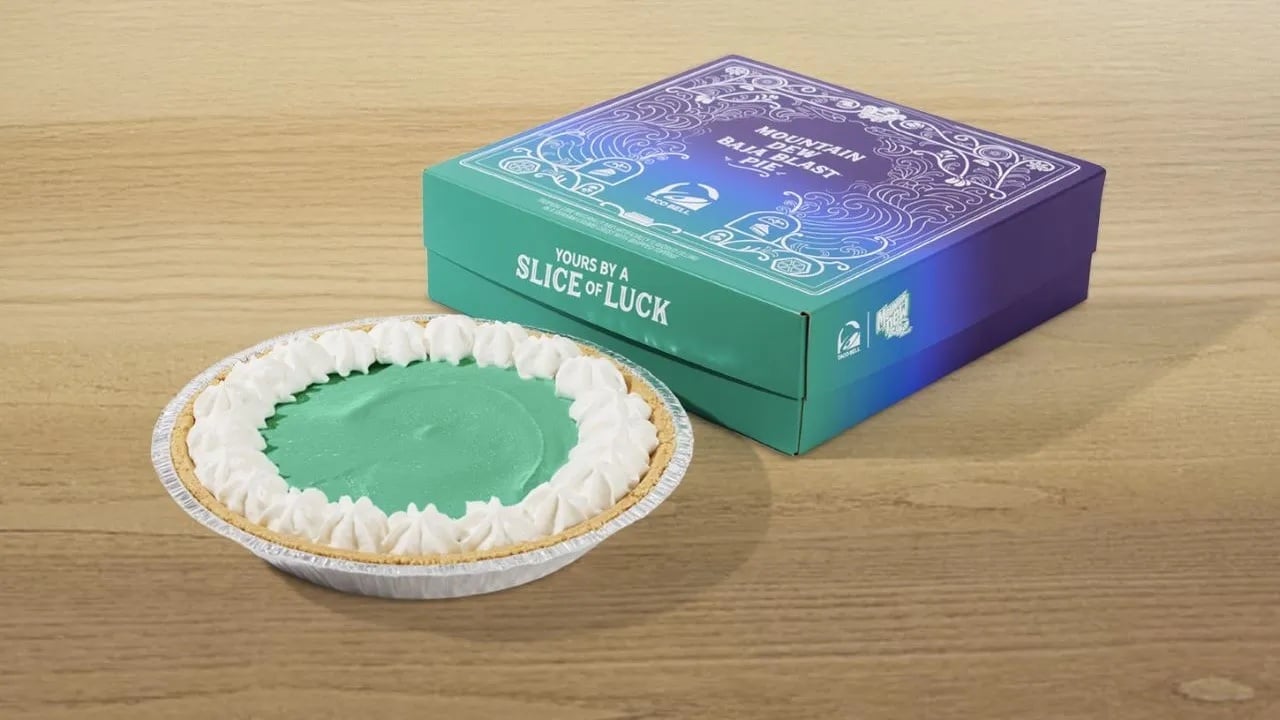 Taco Bell's Baja Blast Pie: A green cream pie with a graham cracker crust and whipped cream border sits on a wooden table next to a blue and purple box labeled "Mountain Dew Baja Blast Pie.