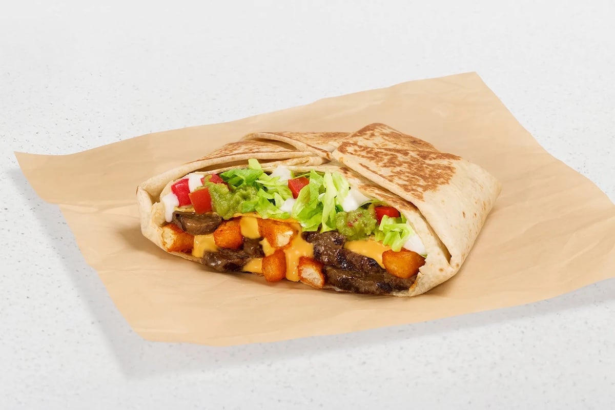 A grilled tortilla wrap cut in half, filled with seasoned beef, diced potatoes, melted cheese, lettuce, diced tomatoes, and sauce, placed on brown parchment paper on a white surface.