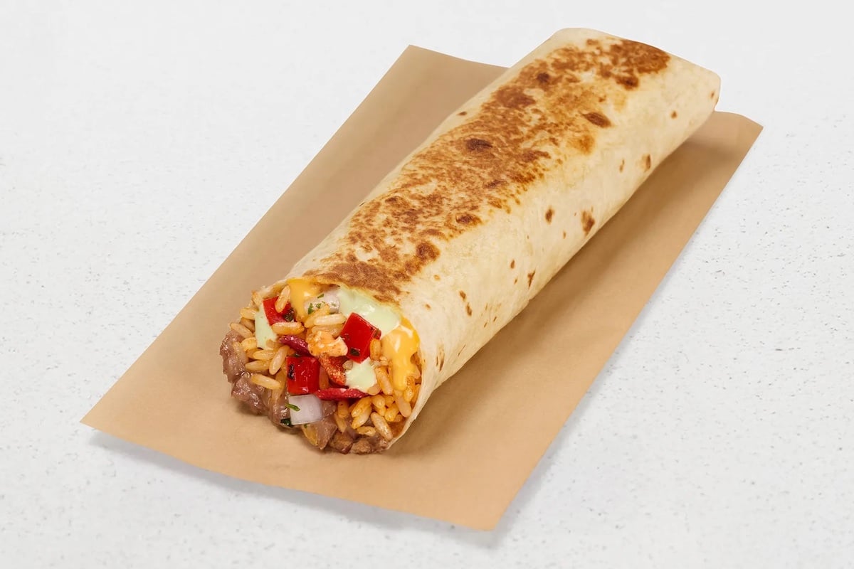 A grilled burrito filled with rice, beans, melted cheese, diced tomatoes, and onions, placed on a sheet of brown paper atop a light speckled surface.