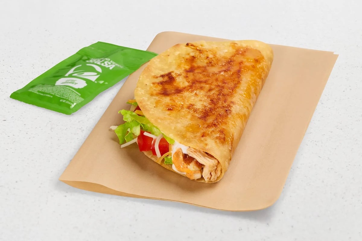 A grilled quesadilla folded in half with visible lettuce, tomato, and shredded cheese, placed on brown paper next to a green packet of salsa on a white surface.