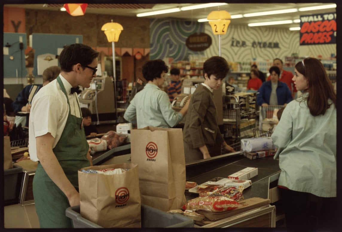 A grocery store checkout lane with a cashier in a green apron scanning items, paper grocery bags on the counter, and customers waiting in line. The background shows more shoppers, products, and colorful signage.