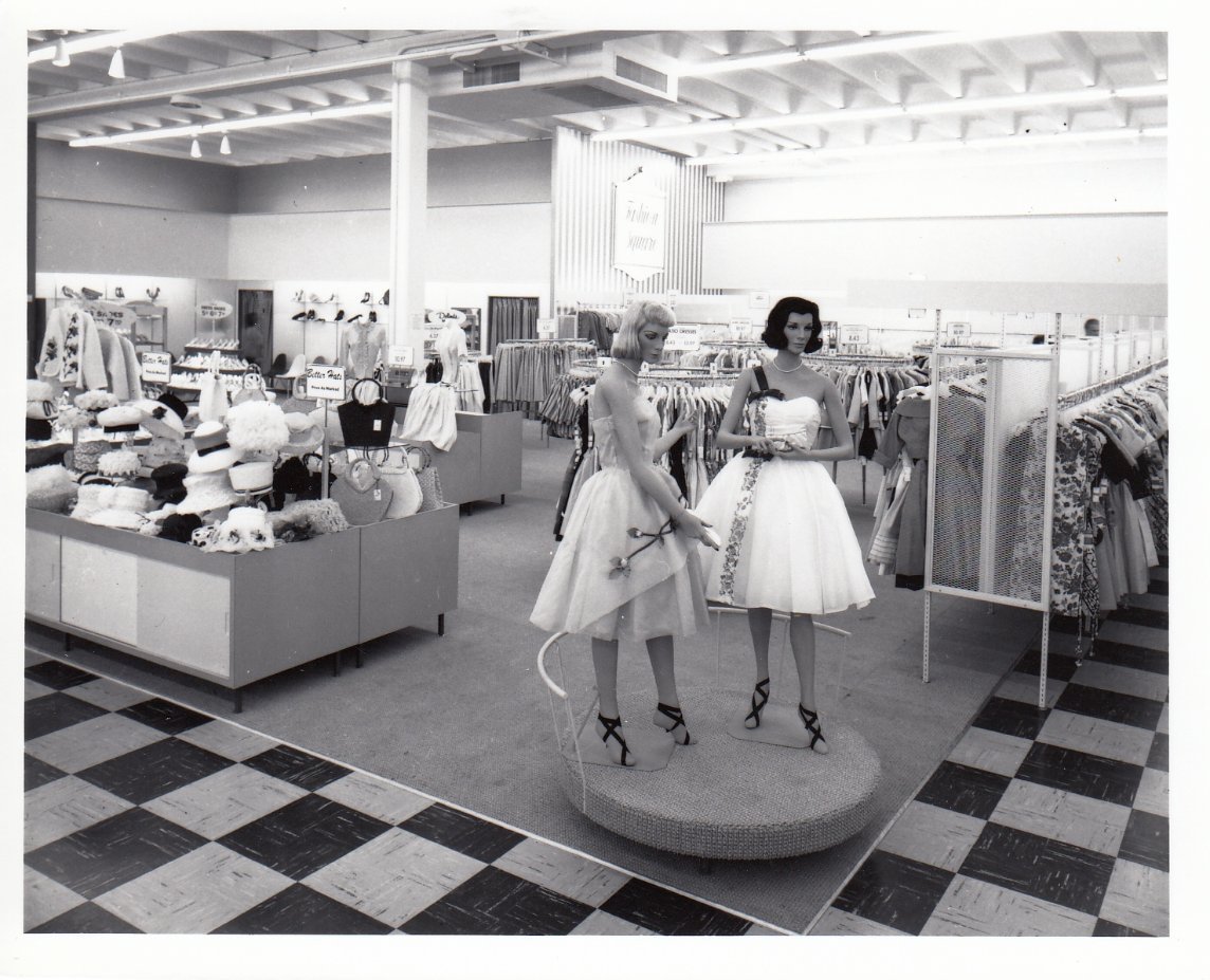 Black-and-white photo of a vintage clothing store with two mannequins in 1950s-style dresses on a circular display. Racks of clothes and hats are visible in the background, and the floor has a checkered pattern.
