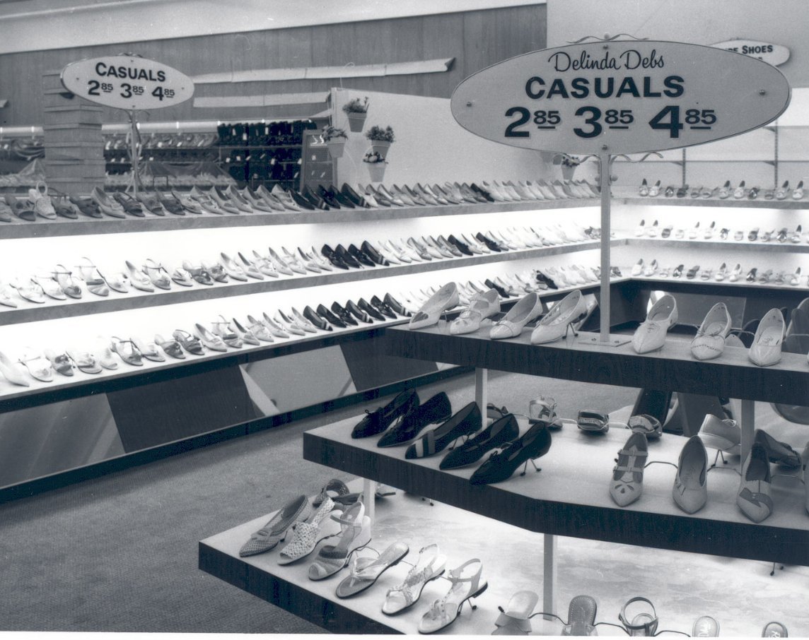 Black-and-white photo of a shoe store with shelves displaying many pairs of women’s casual shoes. A sign reads: "Delinda Debs CASUALS 2.85, 3.85, 4.85." The store interior is brightly lit.