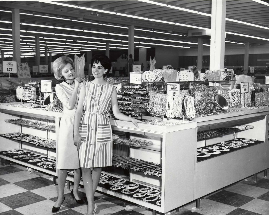 Two women stand by a jewelry display in a store, smiling and looking at each other. The store has checkered floors and shelves filled with necklaces and accessories under bright overhead lights.