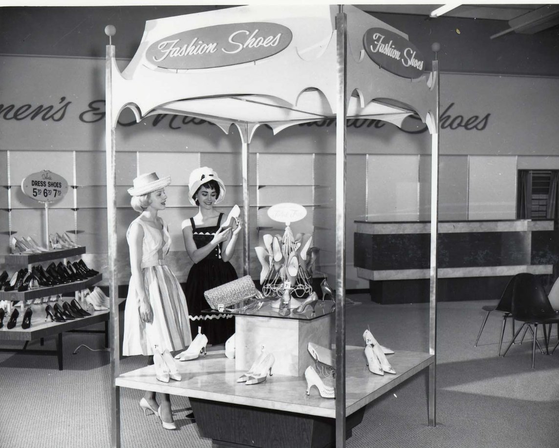 Two women in 1950s-style dresses and hats admire shoes at a "Fashion Shoes" display in a department store. Shelves of shoes and a price sign are visible in the background.