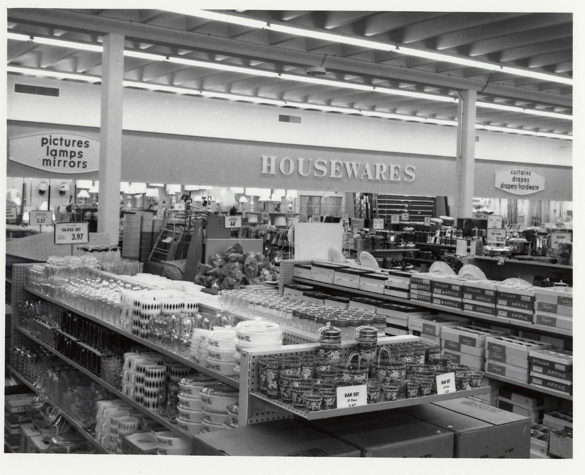 Black and white photo of a store’s housewares section, with shelves displaying glassware, dishes, and storage containers. Signs for “pictures, lamps, mirrors” and “artist, crafts, party hobby” are visible in the background.
