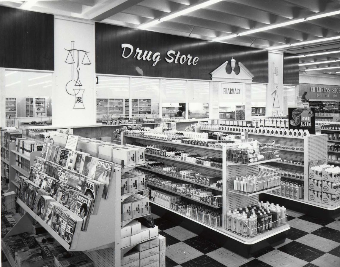 Black-and-white photo of a vintage drug store with shelves of products, a magazine rack, and a "Drug Store" sign above the pharmacy counter. The interior has bright lighting and checkered flooring.