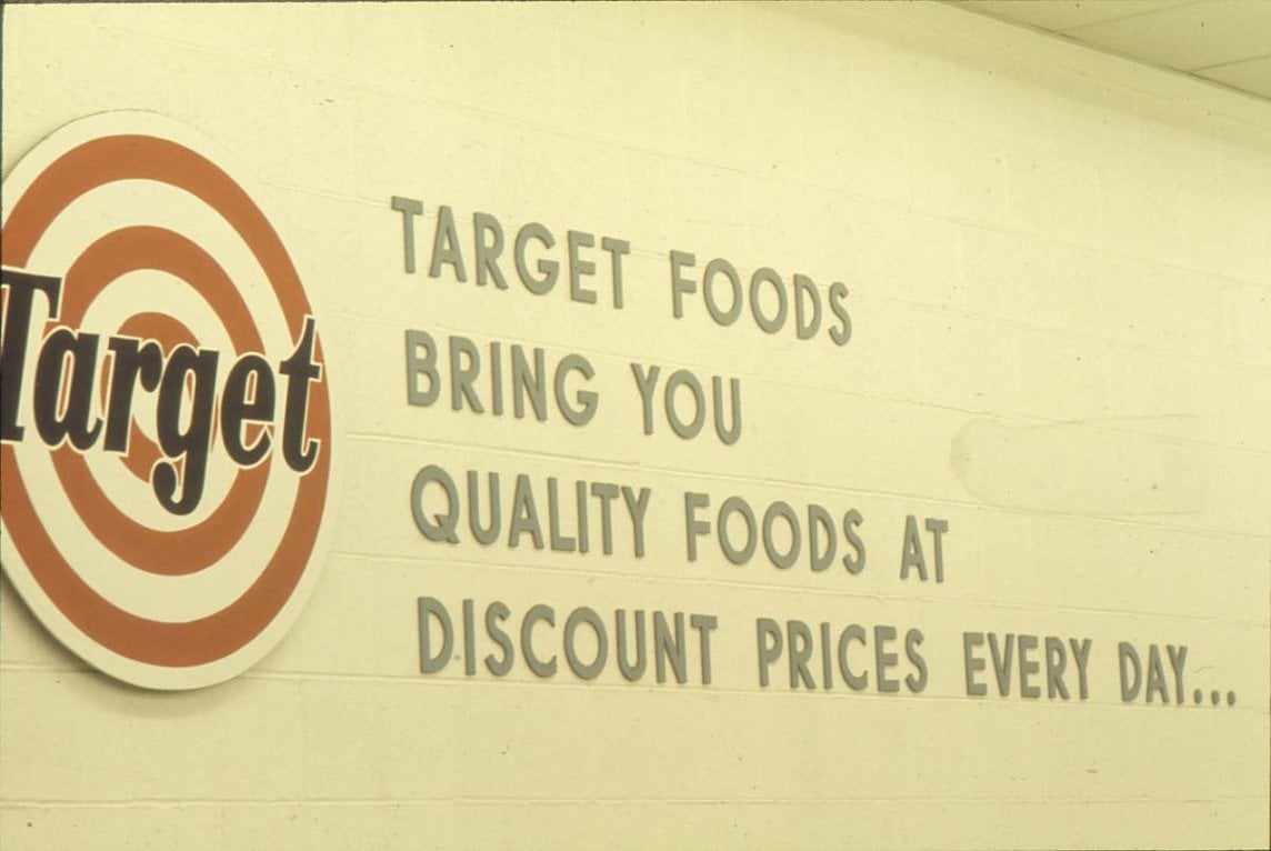 A wall sign reads: “Target Foods bring you quality foods at discount prices every day...” with a Target logo on the left side. The sign is on a light-colored wall.