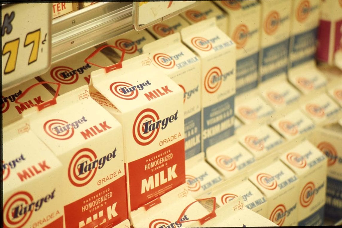 Cartons of Target brand homogenized milk are stacked on a grocery store shelf alongside similar cartons. A yellow price tag is partially visible in the upper left corner.