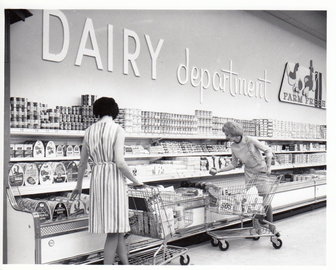 Two women with shopping carts select items from a refrigerated dairy section in a grocery store. Shelves are stocked with various dairy products, and a sign above reads "DAIRY department" with a "FARM FRESH" illustration.