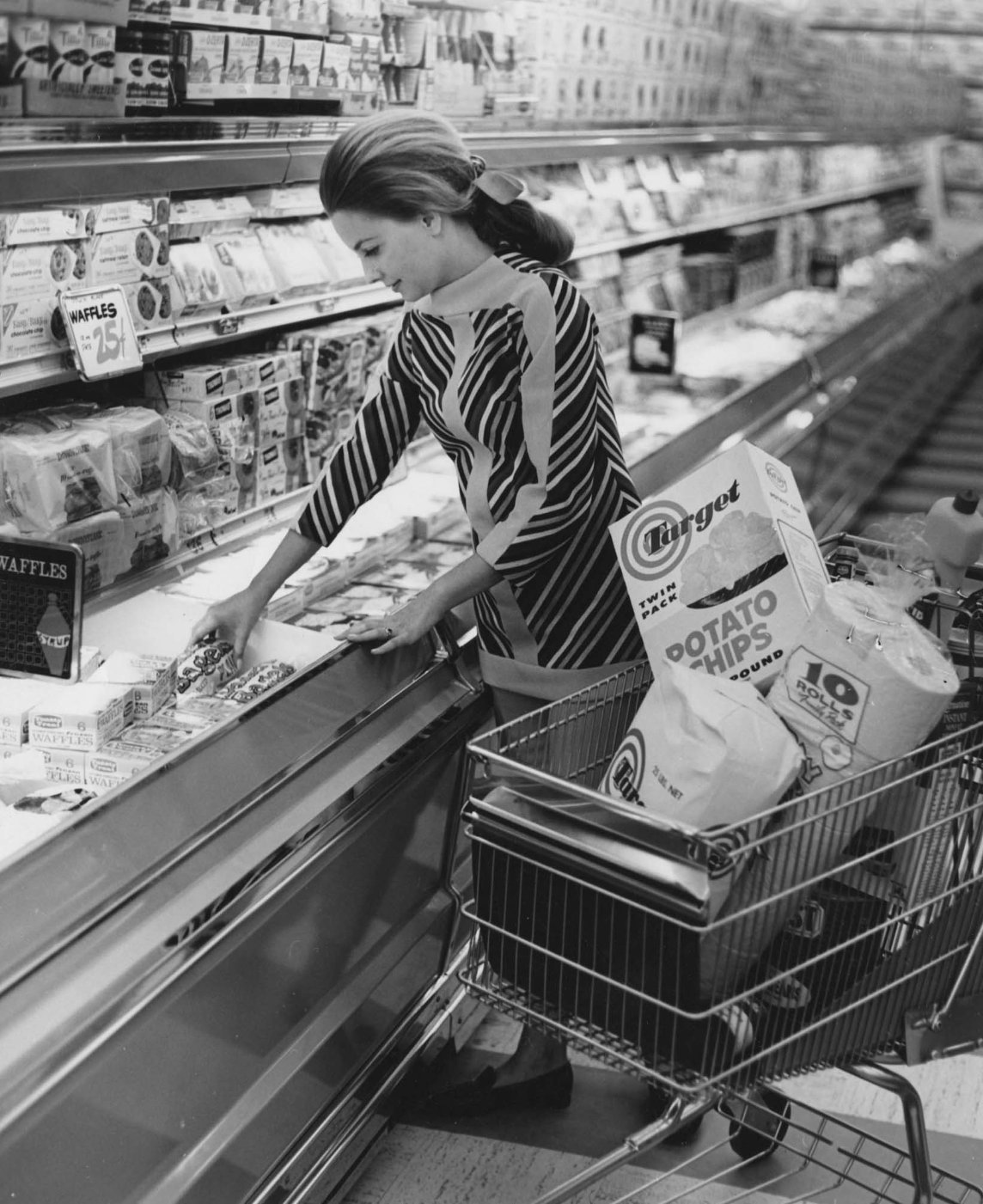 A woman in a striped dress shops in a grocery store, reaching into the freezer section while pushing a cart filled with groceries, including a large bag of potato chips and paper towels.