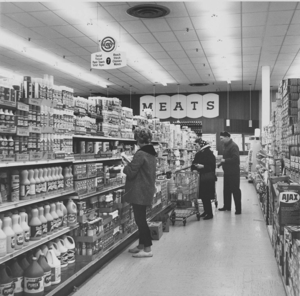 Black and white photo of shoppers in a grocery store aisle lined with shelves of products. A large sign reading "MEATS" hangs at the back. Three people browse or push carts under fluorescent lights.