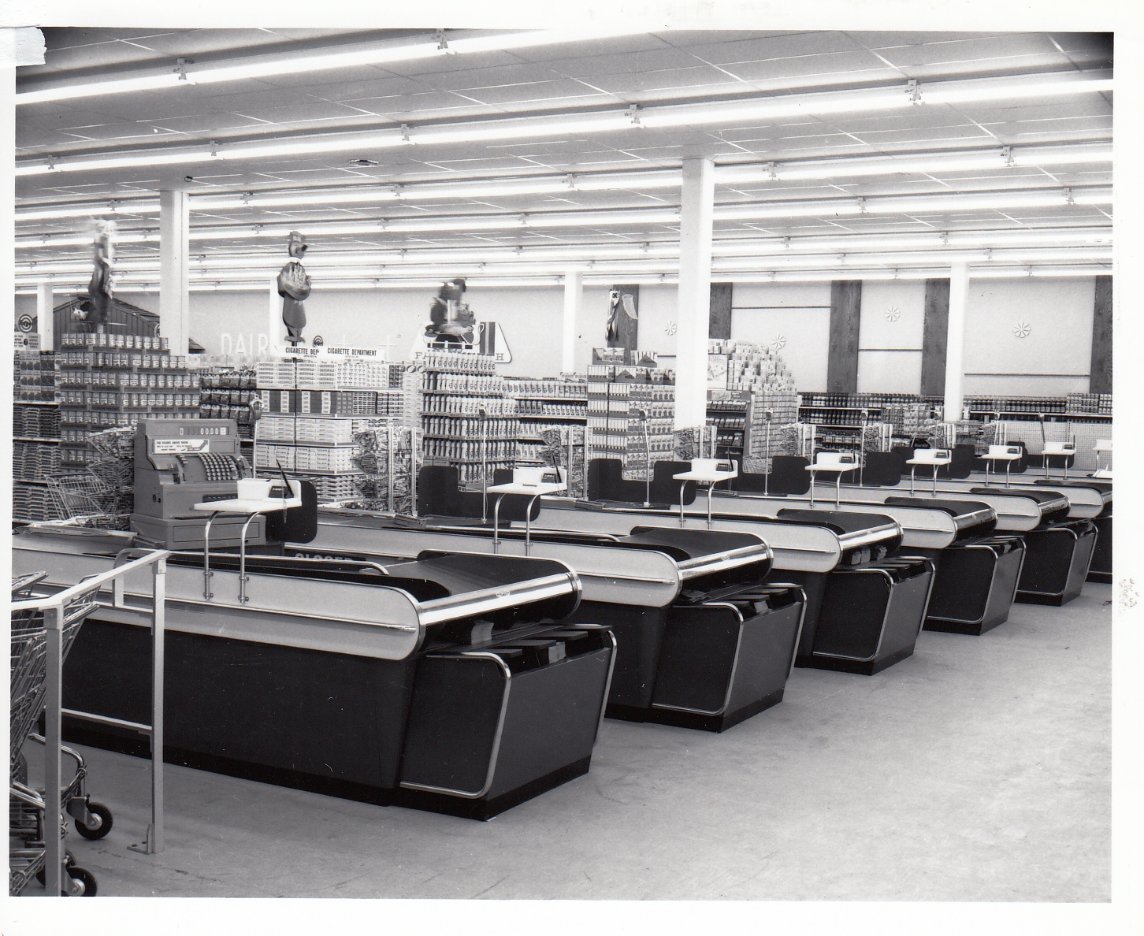 Black and white photo of an empty supermarket checkout area with multiple old-fashioned cash registers, grocery aisles in the background, and a shiny clean floor. No people are present.