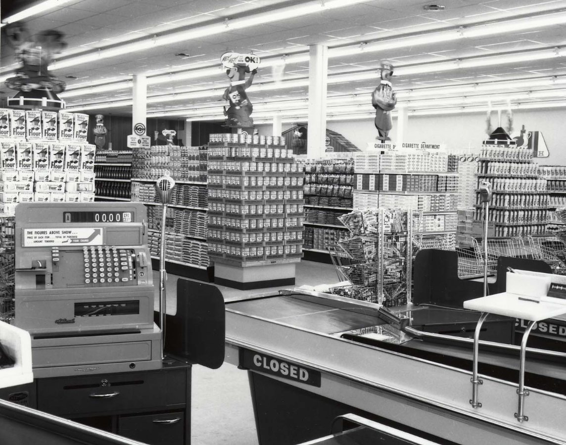 Black-and-white photo of an old-fashioned supermarket with stacked shelves, a vintage cash register, and two checkout lanes marked “CLOSED.” The store is well-lit with ceiling lights and cartoonish displays above aisles.