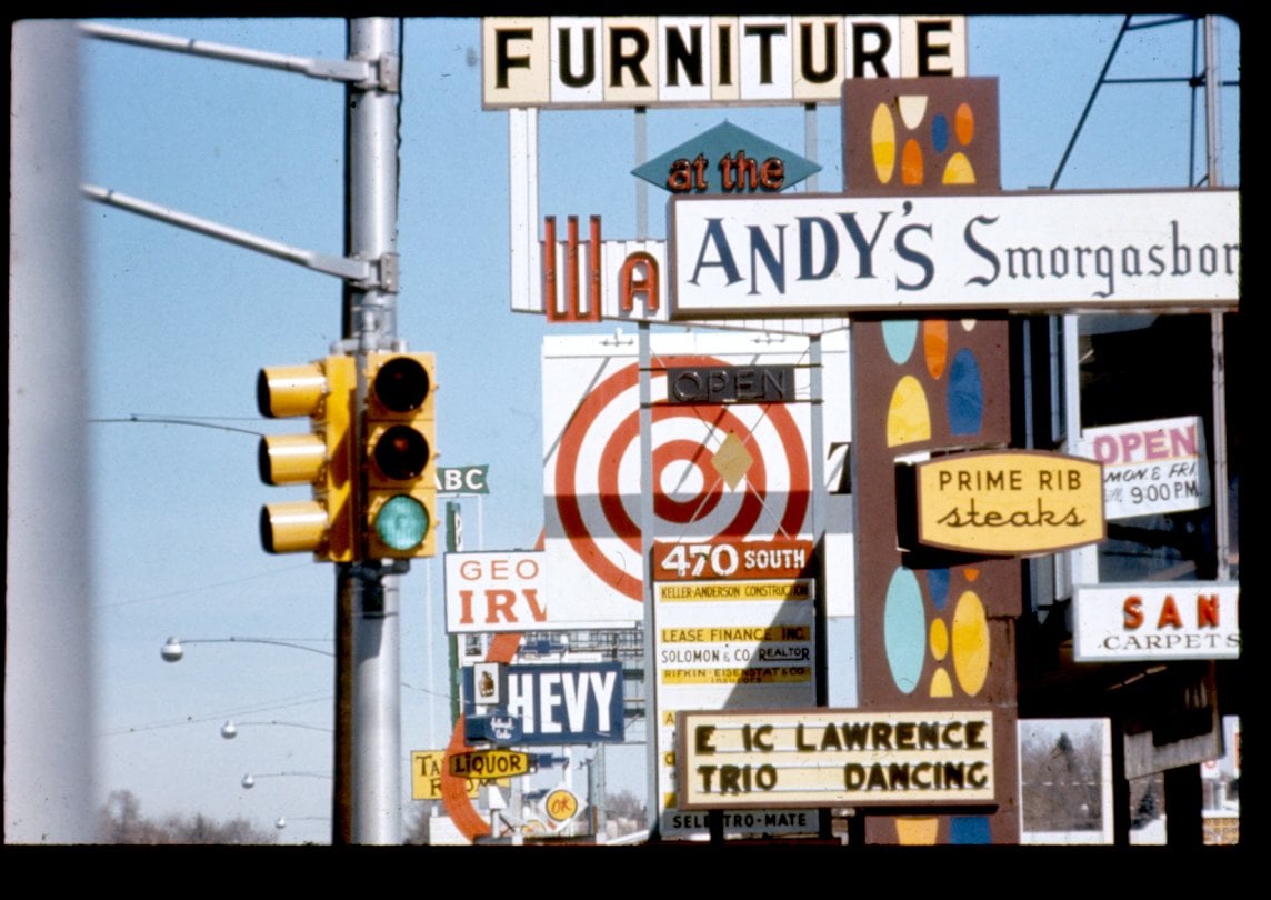 A cluttered street scene filled with colorful, overlapping business signs, including ads for furniture, restaurants, liquor, carpets, and a prime rib steak house. A traffic light and power lines are visible on the left.
